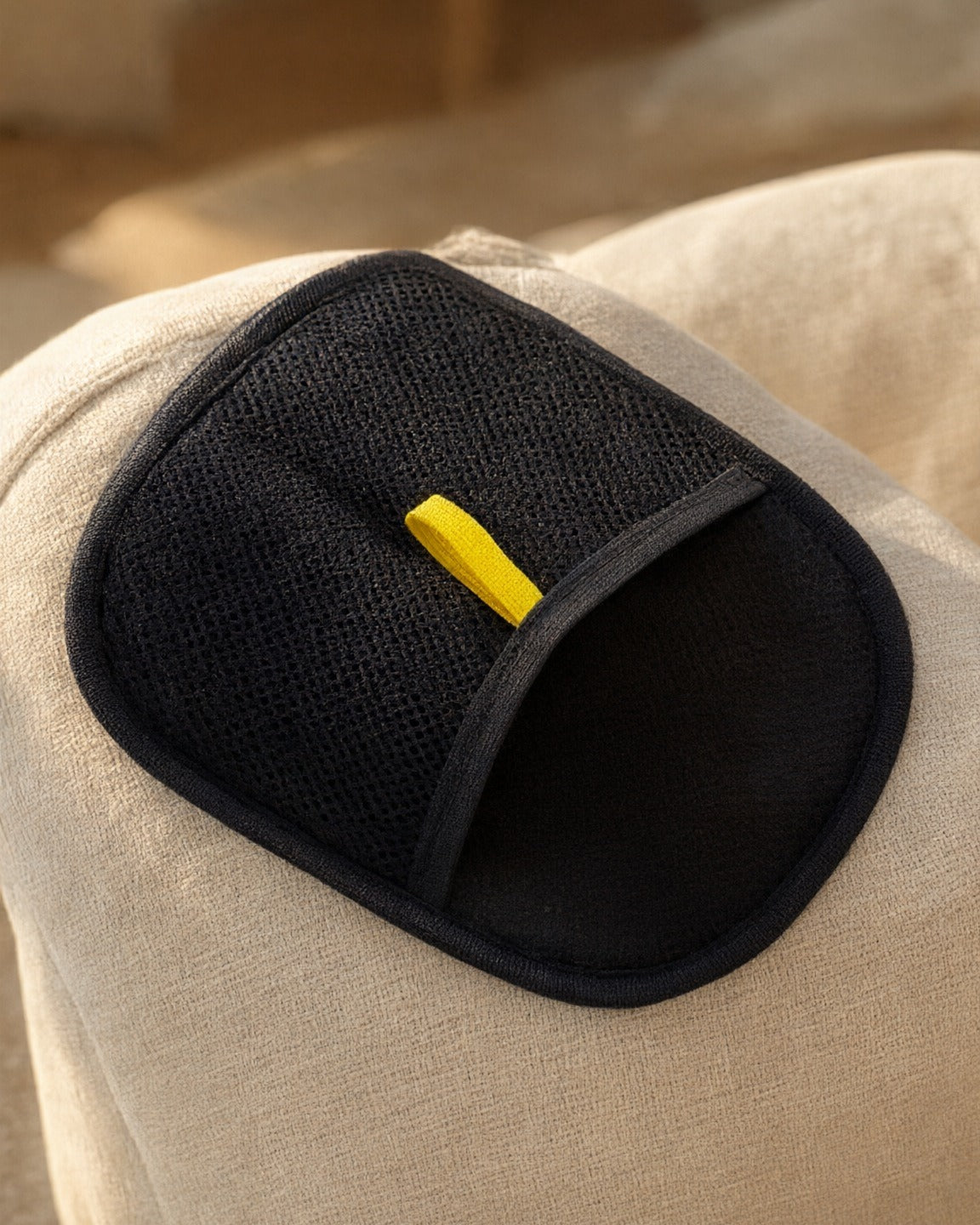 FurSwipe pet hair glove resting on a cream sofa arm, black mesh side with the yellow pull tab