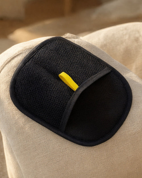 FurSwipe pet hair glove resting on a cream sofa arm, black mesh side with the yellow pull tab